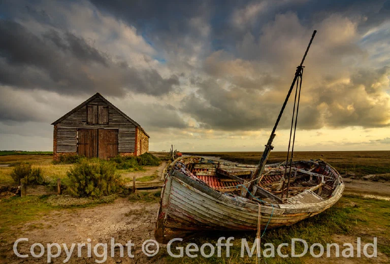 Derelict wooden sailing boat beside an old harbour building under heavy clouds at sunrise