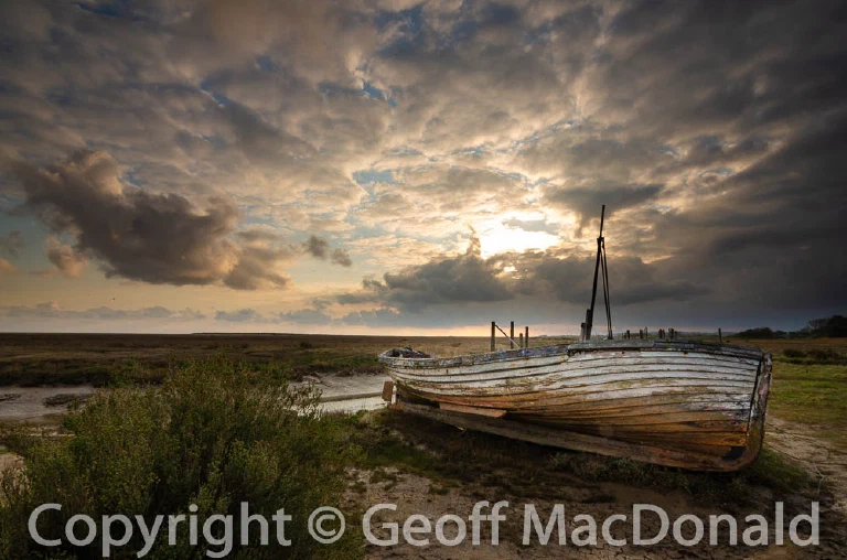 Derelict wooden sailing boat beneath dramatic clouds and the rising sun at Thornham Old Harbour